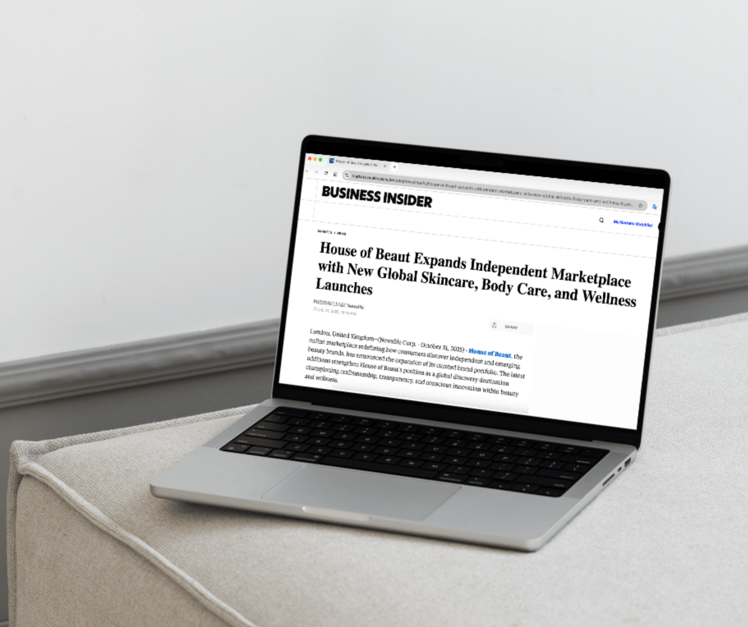 A laptop resting on a light grey fabric surface displays a Business Insider article titled “House of Beaut Expands Independent Marketplace with New Global Skincare, Body Care, and Wellness Launches.” The screen shows the article headline and opening paragraph, highlighting the brand’s growth and new product categories.