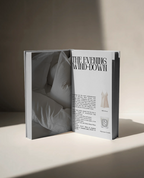 Open book on a neutral surface showing a page titled “The Evening Wind-Down”, featuring soft white bedding imagery and text about evening self-care rituals, including a silk dress and Diptyque candle.